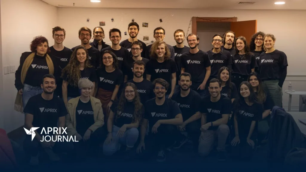 Foto grupal del equipo de Aprix, compuesto por unas treinta personas, todas usando camisetas negras con el logo de la empresa. Están sonriendo y posando en una sala iluminada, decorada con fotos colgadas en la pared. En la esquina inferior izquierda aparece el logo de Aprix Journal.