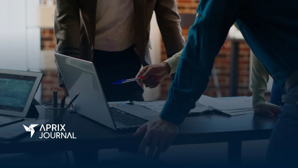Personas colaborando en una mesa de trabajo, revisando documentos y gráficos junto a computadoras portátiles y una tablet con datos financieros en la pantalla. Una mano señala la laptop con un bolígrafo. En la esquina inferior izquierda aparece el logotipo de "Aprix Journal".
