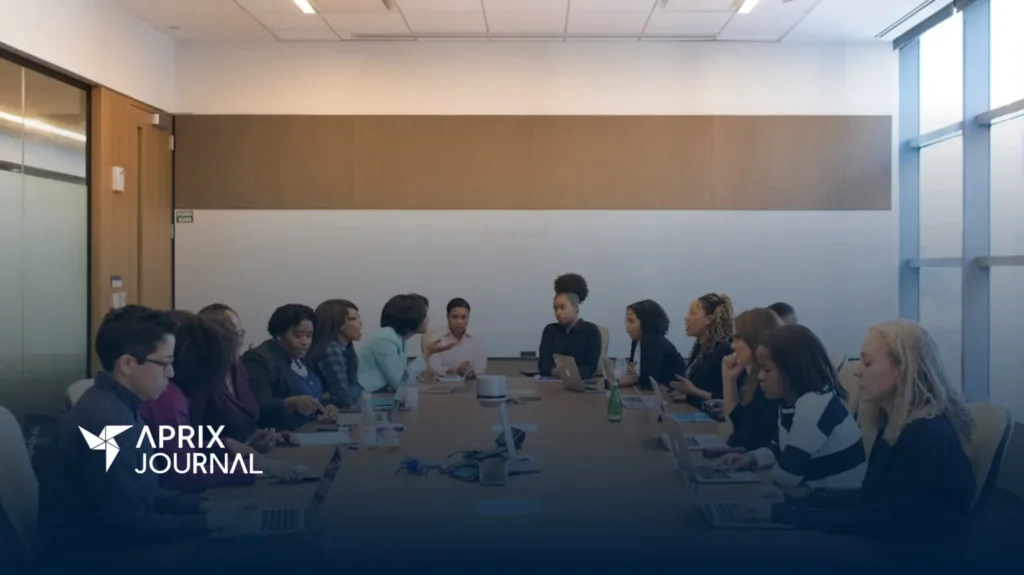 Reunión de trabajo en una sala de conferencias con un grupo diverso de personas, mayormente mujeres, sentadas alrededor de una mesa larga, usando laptops y conversando. En la esquina inferior izquierda se ve el logotipo de "Aprix Journal".